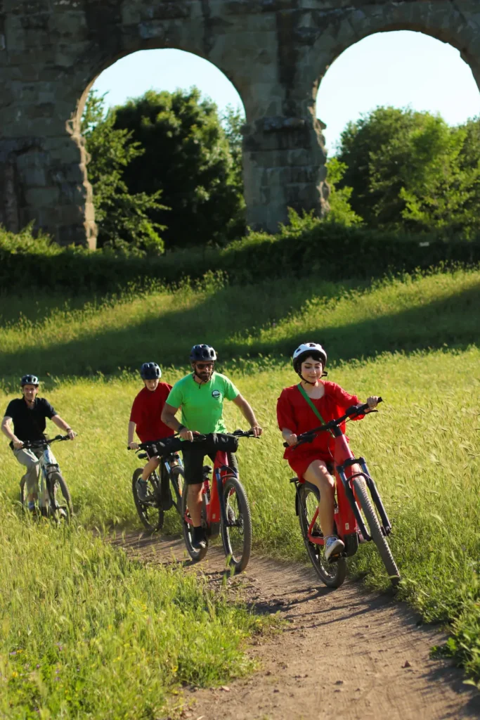 Guide and guests riding e-bikes on a dirt path through a green meadow in front of Roman aqueduct arches in Rome