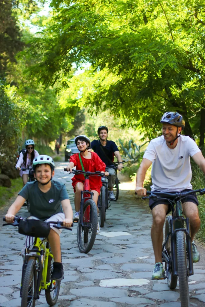 Family riding bikes along the ancient cobblestone Appian Way in Rome, shaded by green trees